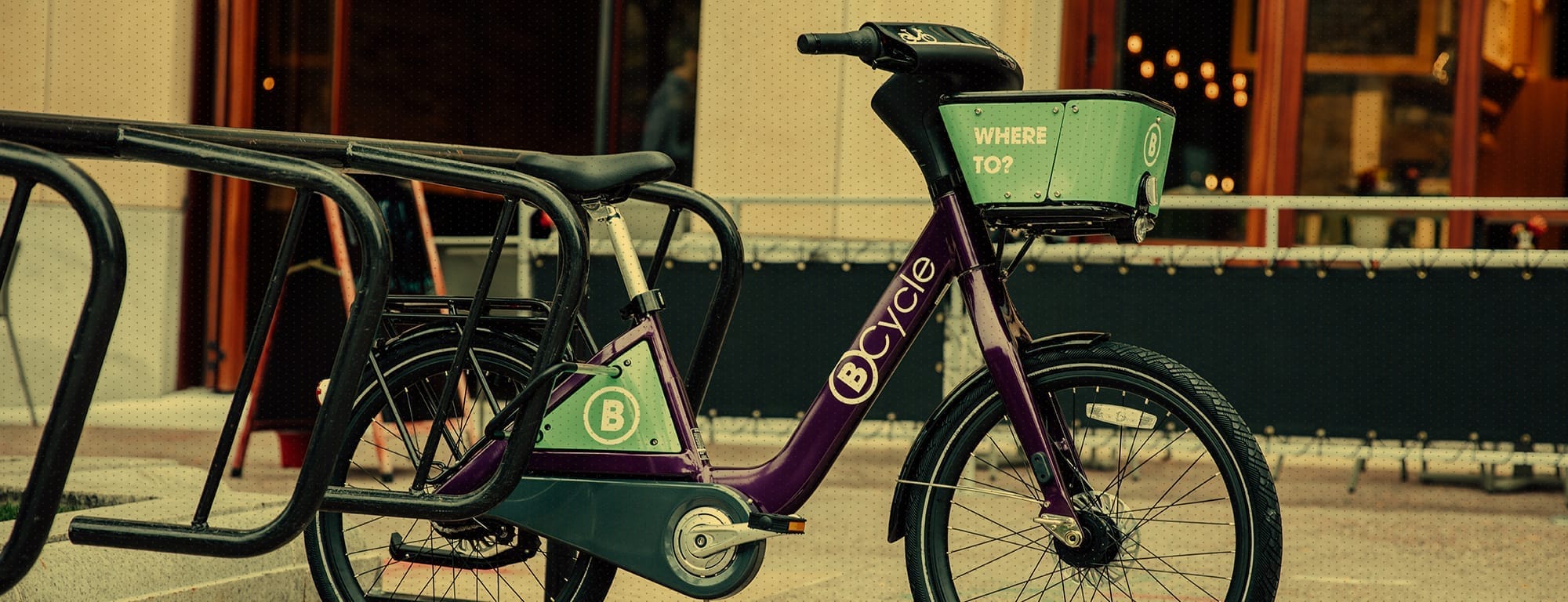 Ein lilafarbenes Leihfahrrad der Marke BCycle mit einem hellgrünen Korb steht an einem Fahrradständer aus Metall auf dem Bürgersteig vor einem Gebäude.