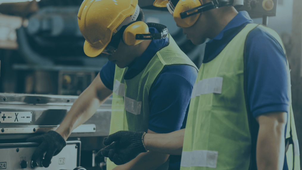 Two industrial workers with yellow vests and hard hats working on a machine