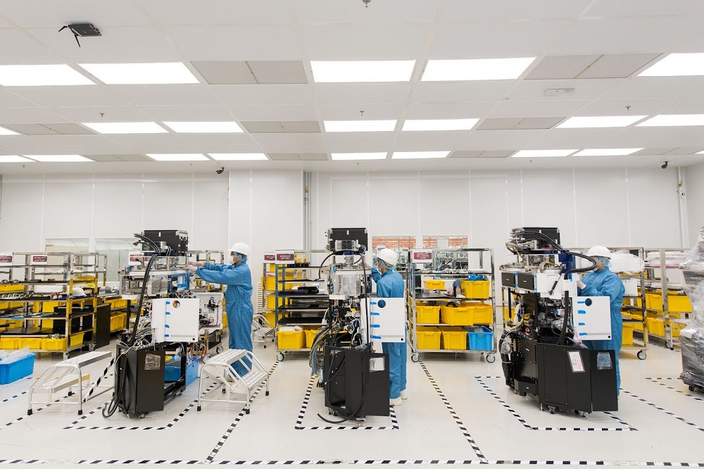 A clean, well-lit manufacturing facility with three workers in blue jumpsuits and white hard hats. They are each working on a piece of large, complex machinery with yellow storage bins and metal racks visible in the background. 
