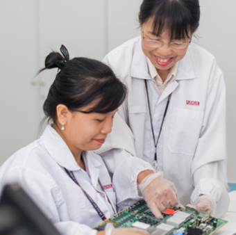 Two women in white lab coats with a Plexus logo and gloves smiling while working together on a complex green circuit board.