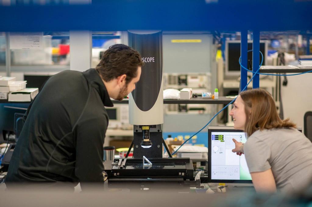 A man and a woman in a high-tech laboratory or manufacturing setting are collaborating over a piece of precision machinery. The man, on the left, leans in to inspect a small component placed on the stage of a digital measuring microscope. The woman, on the right, smiles while pointing to data on a computer monitor that displays the machine's interface. The background shows a professional workspace filled with electronics and testing equipment.