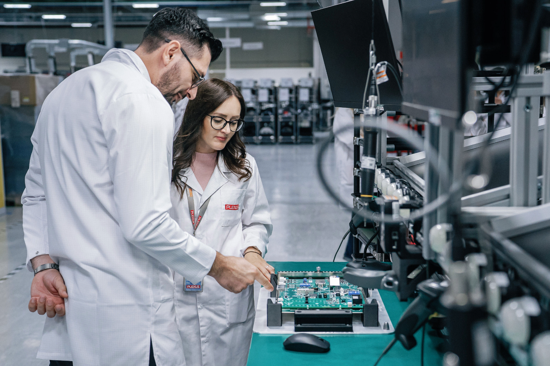Two Plexus engineering team members at the Oradea, Romania facility meticulously inspecting a complex printed circuit board assembly (PCBA). Both technicians wear required ESD-safe anti-static smocks and protective gear while performing visual quality control and verify component placement accuracy on the high-mix line.